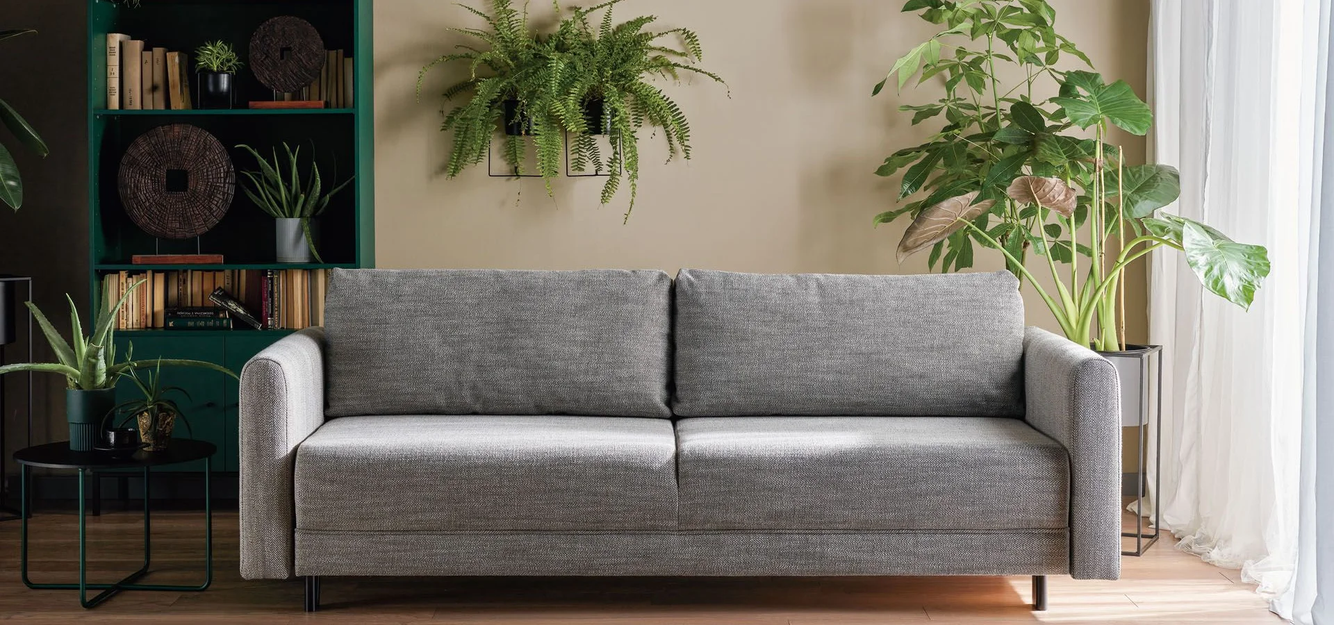 Szara sofa w otoczeniu zielonych roślin i regału z książkami.